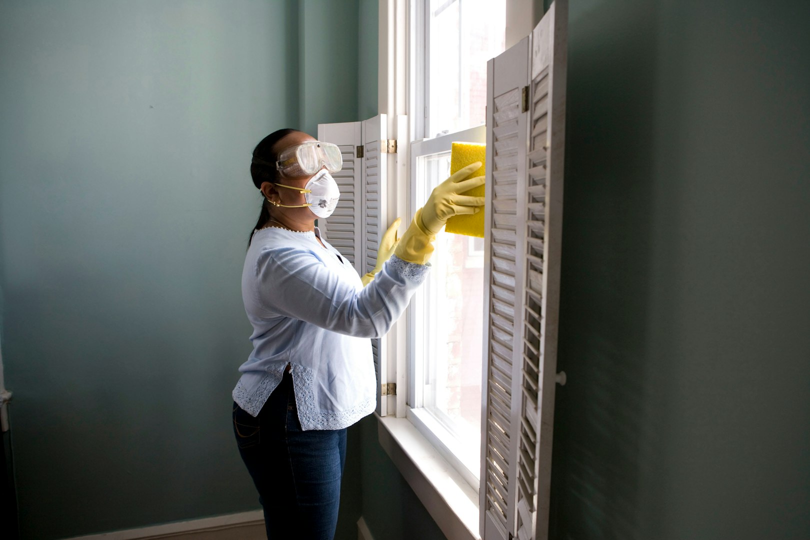 A woman wearing yellow rubber gloves is cleaning a shelf in a bright, modern living space. She is arranging decorative items and plants on the shelf, while a man sits casually on a couch in the background.