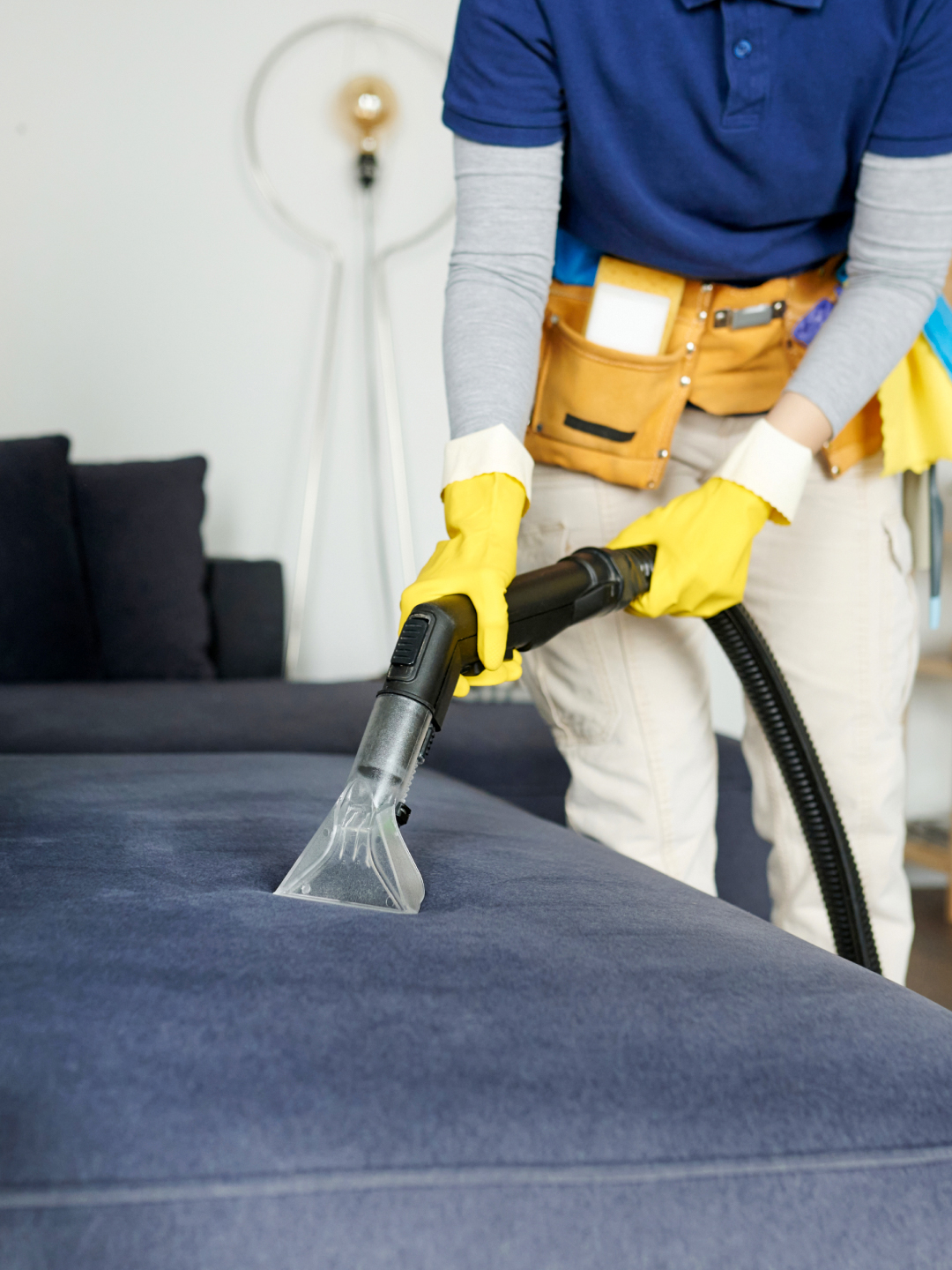 A person wearing yellow gloves uses a vacuum cleaner attachment to clean a dark blue upholstered couch. The individual is wearing a blue shirt and a tool belt, and there is a lamp in the background.