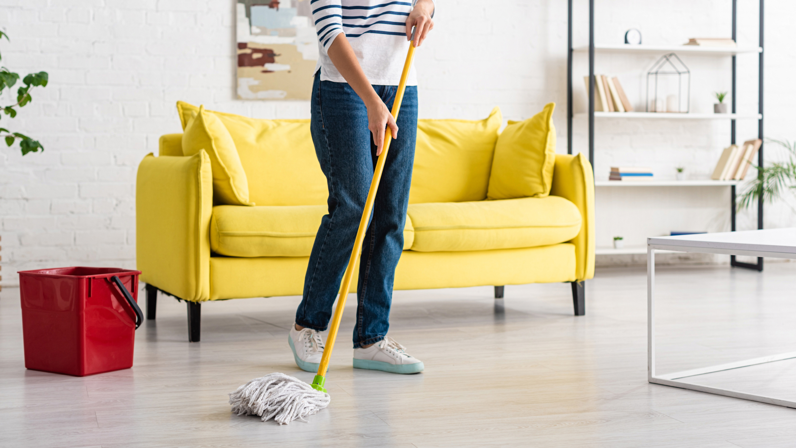 A person is mopping a light-colored floor in a spacious living room with a yellow couch, a red bucket nearby, and white shelves with books and decor in the background.